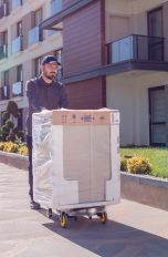 Eine Person schiebt eine eingepackte Waschmaschine vor einem Wohnkomplex.